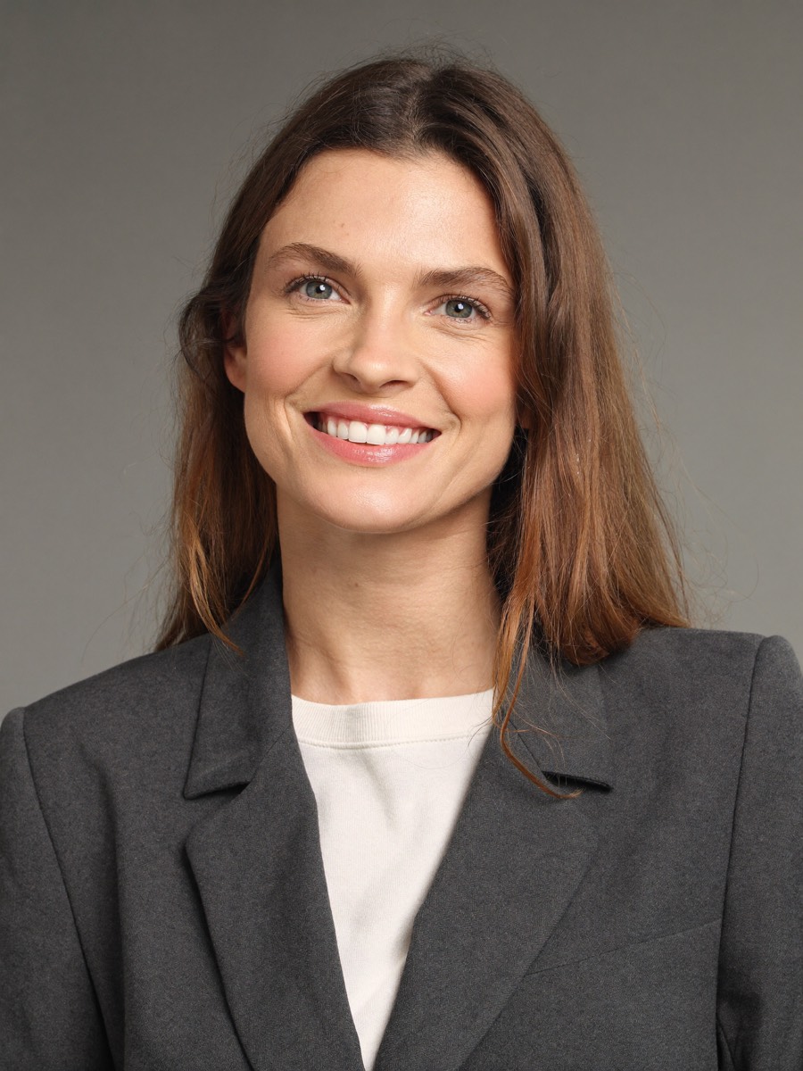 A smiling woman in a charcoal blazer against a plain grey studio backdrop