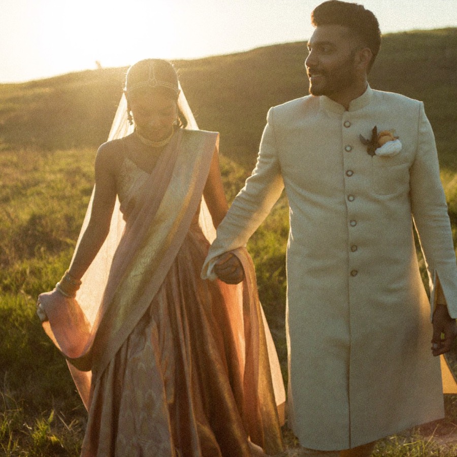 A bride and groom walking hand in hand through a meadow at golden hour